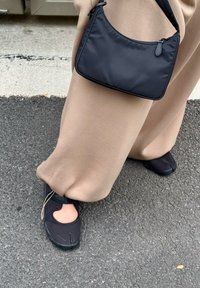 Chaussures en tissu noir avec design découpé, associées à un pantalon large beige et un petit sac à épaule noir, reposant sur une surface pavée sombre.