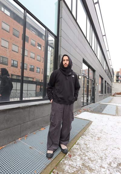 Sudadera negra con capucha, pantalones cargo grises y holgados con bolsillos, zapatos negros. De pie sobre una rejilla de metal gris junto a un edificio moderno con reflejos.