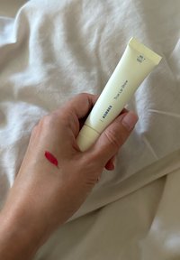 Käsi pitää Korres True Lip Shine -putkiloa, jossa on punaisen huulikiillon kokeilu iholla, pehmeän valkoisen kankaan taustaa vasten.