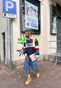 Χρωματιστό ριγέ μπουφάν με το λογότυπο "TOMMY", φαρδιά τζιν σορτς, κίτρινες μπότες και ακουστικά, κρατώντας ένα περιοδικό δίπλα σε έναν τοίχο με γκράφιτι.
