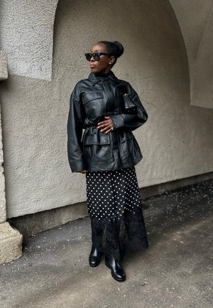 Mujer con chaqueta de cuero negra, falda de lunares con borde de encaje, botas negras, gafas de sol, de pie bajo un arco sobre un suelo pavimentado.