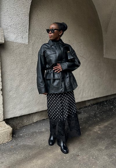 Mujer con chaqueta de cuero negra, falda de lunares con borde de encaje, botas negras, gafas de sol, de pie bajo un arco sobre un suelo pavimentado.