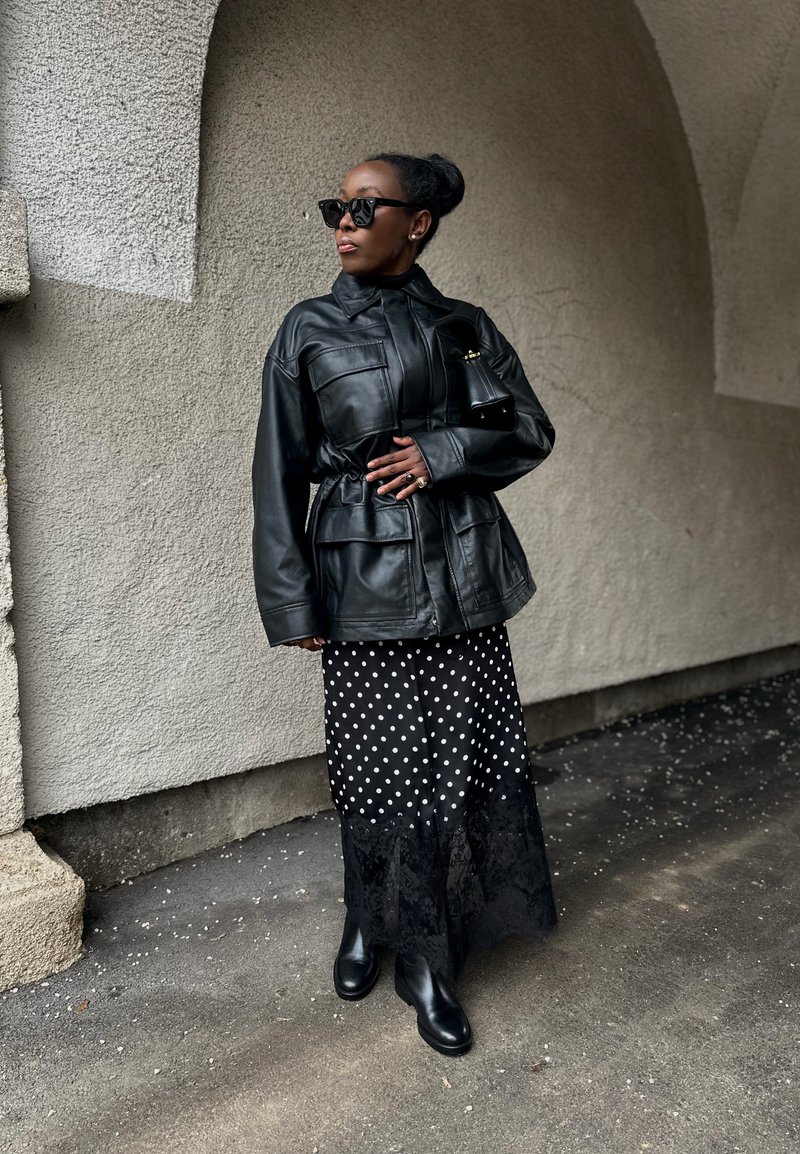 Femme portant une veste en cuir noire, une jupe à pois avec un ourlet en dentelle, des bottes noires, des lunettes de soleil, debout sous une arche sur un sol pavé.