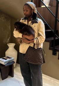 Vevd, mørkebrunt skinntote med en strukturert form og to flettede håndtak. Beige skjorte med krage, holder en fluffy brun hund.