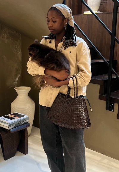 Bolso de piel trenzada de color marrón oscuro con forma estructurada y dos asas trenzadas. Camisa beige con cuello, sosteniendo un perro marrón esponjoso.