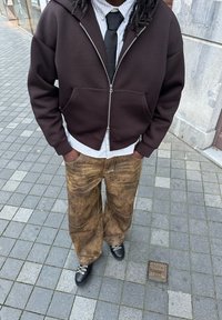 Brauner Reißverschluss-Hoodie über einem gestreiften Shirt, kombiniert mit weiten beigefarbenen Hosen. Schwarze Schuhe mit weißen Schnürsenkeln, auf einem grauen Pflaster.