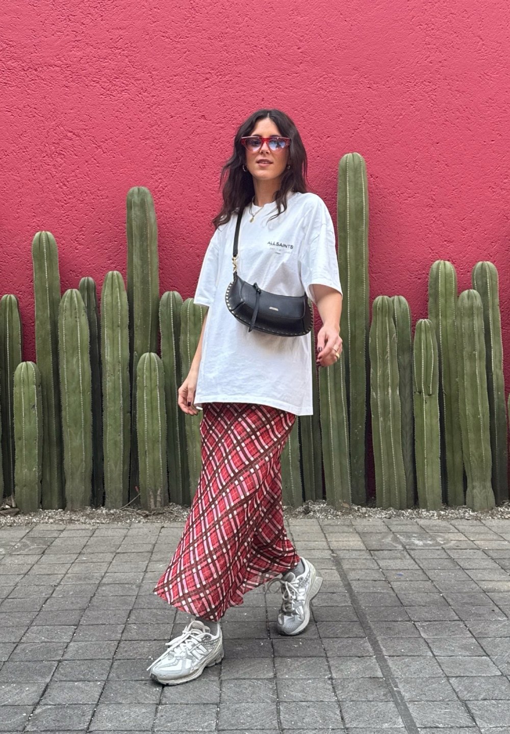 T-shirt oversized branca com o texto "ALLSAINTS", saia maxi xadrez vermelha, mala de crossbody preta e ténis cinzentos. O fundo apresenta cactos verdes altos.