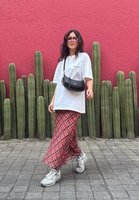 T-shirt oversized branca com o texto "ALLSAINTS", saia maxi xadrez vermelha, mala de crossbody preta e ténis cinzentos. O fundo apresenta cactos verdes altos.