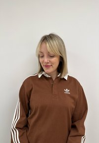 Polo de culoare marron cu mânecă lungă, cu guler alb și trei dungi albe pe mâneci. Are un logo adidas brodat pe piept.