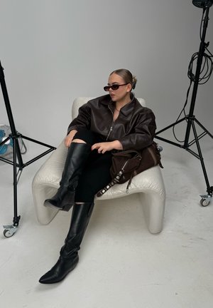 Chaqueta de cuero marrón de gran tamaño, pantalones ajustados negros, botas negras hasta la rodilla y gafas de sol, sentado en una silla blanca curvada.