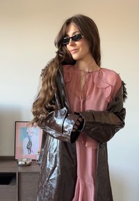 Hnedý lesklý dlhý kabát navrchu ružovej blúzky s volánikovými detailmi, s dlhými rukávmi a výrezom na krku.