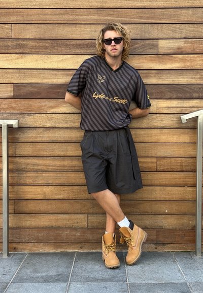 Camisa de manga corta a rayas negras con logo dorado, combinada con pantalones cortos negros hasta la rodilla y botas de trabajo marrones con cordones amarillos, calcetines blancos.