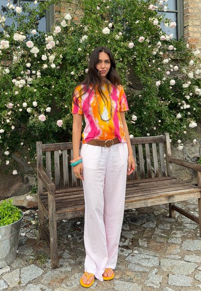 Mujer con cabello largo y oscuro que lleva una camiseta teñida en rosa, naranja y blanco, pantalones blancos, sandalias amarillas, de pie junto a un banco de madera y un arbusto en flor.