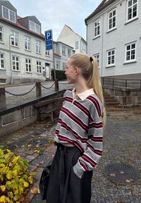 Pasiasty, szary i burgundowy longsleeve polo z jasnym kołnierzykiem, zestawiony z wysokim stanem ciemnych spodni. Tło to brukowana ulica.