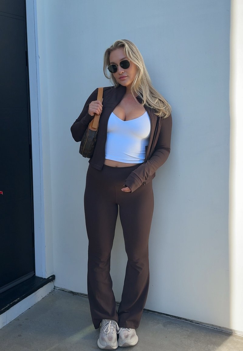 Mujer con chaqueta y pantalones marrón oscuro, con top corto blanco y zapatillas deportivas, de pie contra una pared clara, sosteniendo un bolso de hombro con gafas de sol.