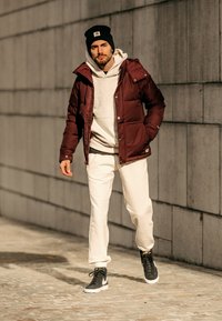 Burgunderfarget polstret jakke med hette over en lys grå hettegenser, hvite joggebukser og svarte joggesko. Står på steinbelagt fortau mot en vegg.