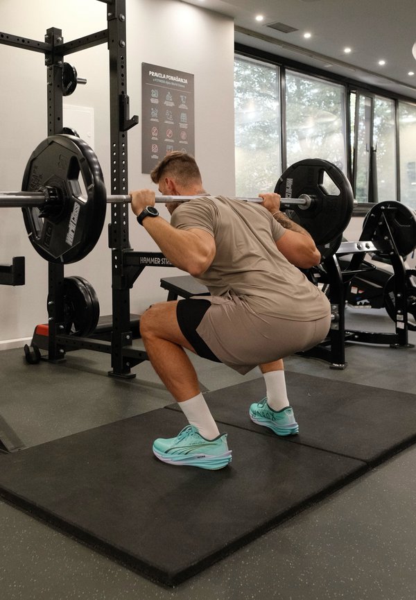 Un hombre en un gimnasio hace una sentadilla profunda con una barra cargada sobre los hombros.