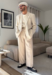 Fato de linho leve em bege com um casaco de duas fileiras de botões, combinado com uma camisa branca canelada e sapatos pretos. Os acessórios incluem uma capa branca e óculos de sol.