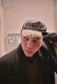 Jonge man met een versleten baseballpet, een donkere jas en handschoenen, die zijn pet rechtzet terwijl hij binnenshuis naast brievenbussen opzij kijkt.