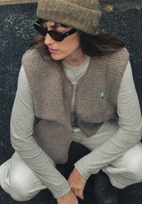 Brązowa kamizelka z polaru z guzikami z przodu, szara koszula z długim rękawem i beżowe spodnie. Na sobie ciemne okulary przeciwsłoneczne i zieloną dzianą czapkę.