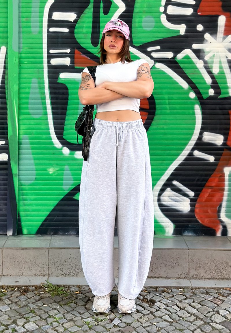 Jeune femme avec tatouages porte une casquette rose, un crop top blanc et un pantalon gris ample, debout les bras croisés devant un mur de graffiti vert et noir.