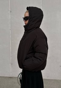 Casaco puffer isolado preto com capuz, apresentando uma forma larga e um colarinho alto. O tecido parece liso e mate.