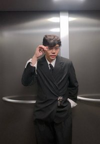 Costume noir à rayures avec larges revers, chemise blanche et cravate noire. Le modèle porte des lunettes rondes et pose dans un ascenseur. Quincaillerie argentée visible.