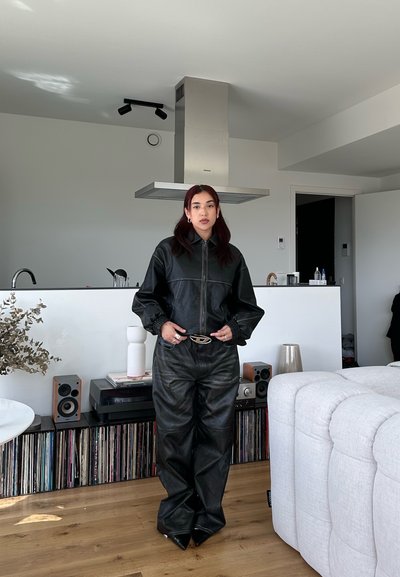 Chaqueta de cuero negra con cremallera frontal, combinada con pantalones de cuero negros sueltos. Incluye una hebilla de cinturón con gráfico. Fondo neutro con mobiliario.