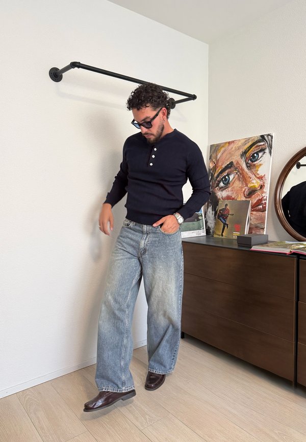 Mand med krøllet hår og solbriller iført en sort bluse med lange ærmer og vide jeans, der står ved siden af en kommode med kunstværker og et rundt spejl.
