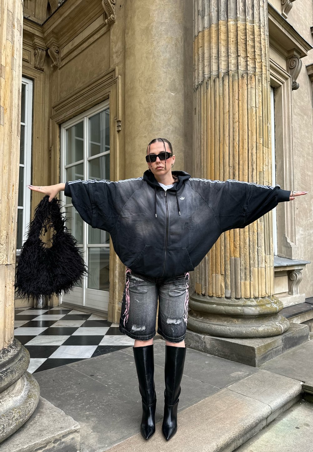 Hanorac negru supradimensionat cu glugă și mâneci largi, pantaloni scurți gri din denim uzat, cizme negre cu toc și până pe coapsă și o geantă neagră pufoasă.