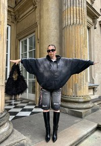 Hanorac negru supradimensionat cu glugă și mâneci largi, pantaloni scurți gri din denim uzat, cizme negre cu toc și până pe coapsă și o geantă neagră pufoasă.