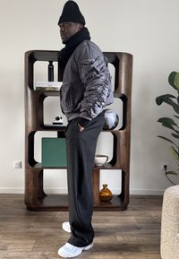 Homme portant un bonnet noir en tricot, une écharpe, une veste aviateur grise, un pantalon noir et des baskets blanches, debout de profil près d'une étagère en bois avec des objets décoratifs.