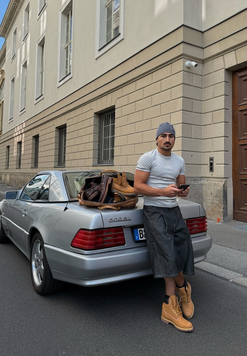 Hopeanvärinen Mercedes-Benz 500 SL parkkeerattuna kadulle. Harmaapaitainen ja sinishortseihin pukeutunut mies nojautuu autoa vasten, jalassaan vaaleanruskeat saappaat. Laukkuja auton takakontilla.