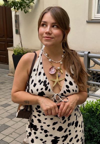 Vestido blanco con manchas negras, con un diseño de lazo en el frente. Collares con estrellas de mar y conchas doradas sobre perlitas y cadena. Bolso marrón.