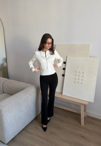 Femme ajustant un cardigan blanc boutonné, portant un pantalon noir et des lunettes, debout près d'un canapé beige et d'une œuvre d'art minimaliste texturée sur un banc en bois.