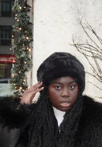 Zwarte nep-bontmuts gedragen bij een zwarte bontjas. De achtergrond is versierd met groenblijvende slingers en zacht licht van decoratieve lampjes.