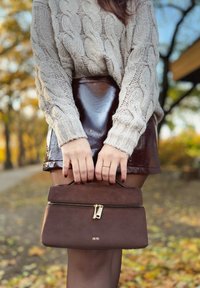 Braune Wildlederhandtasche mit Reißverschluss und goldenen Beschlägen, von Händen vor einem glänzenden burgunderfarbenen Rock und einem cremefarbenen Kabelstrickpullover gehalten.