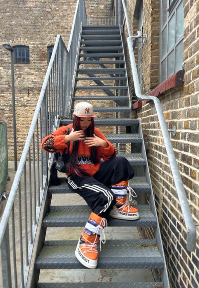 La modelo se sienta en unas escaleras de metal vistiendo un suéter de lana naranja, pantalones negros con rayas blancas y zapatillas Moon Boot naranjas con patrones de rayos.
