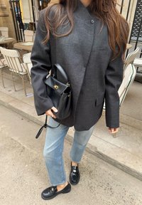 Pelēks struktūrēts žakete ar mandarīna apkakli, kopā ar gaiši zilām taisnām džinsām un melnām loafer apaviem. Melna rokassoma ar zelta akcentiem.