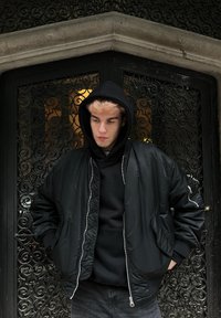 Jeune personne vêtue d'un sweat à capuche noir et d'une veste bomber, les mains dans les poches, se tenant devant une grille en fer forgé ornée.