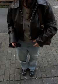 Must musta nahkjakki rullitud varrukatega, hall nööbiga särk, helesinised häiritud teksad ja mustad nupukesed kingad, hoides päikeseprille.