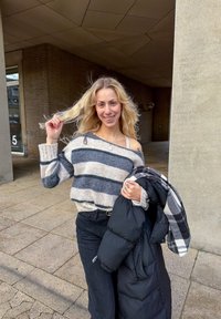 Pulover tricotat cu dungi în gri, bej și negru, cu croială lejeră. Geacă de puf neagră așezată pe un braț. Pantaloni negri. Zâmbind.