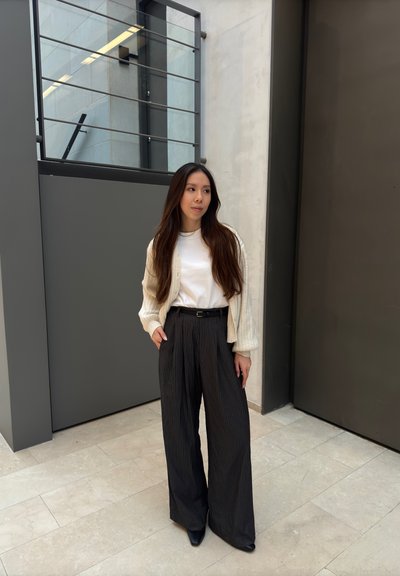 Amplios pantalones a rayas negros combinados con una blusa blanca y un cárdigan texturizado color crema. De pie sobre un suelo de piedra de color claro.