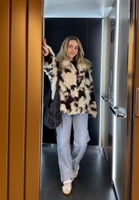 Műszőrme kabát fekete és krémszínű mintával, világoskék bővülő szárú farmer, fehér tornacipő. Fekete táska a kar alatt tarkónál. Lift környezetben.