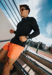 Mann in schwarzem Langarmshirt und orangefarbenen Shorts, der an einer Sportbahn unter einem blauen Himmel die Außentreppen joggt.