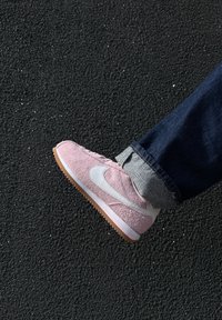 Rosa Wildleder Sneaker mit weißen Akzenten und einer beigen Gummisohle, mit abgerundeter Zehe und strukturierter Oberfläche, vor einem dunklen Hintergrund.