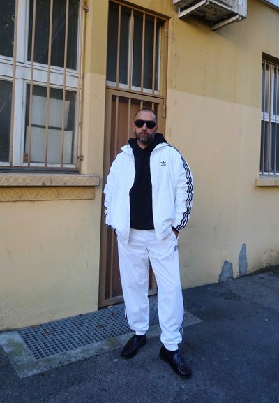 Hombre vistiendo un chándal blanco de Adidas y una sudadera negra, de pie con las manos en los bolsillos frente a un edificio beige con ventanas enrejadas.