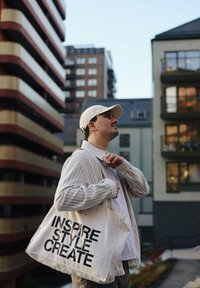 Geantă de pânză cu text negru "ÎNDRUMĂ STIL CREEAZĂ." Bărbat îmbrăcat cu un cardigan bej deschis și o șapcă, stând în fața unei clădiri moderne.