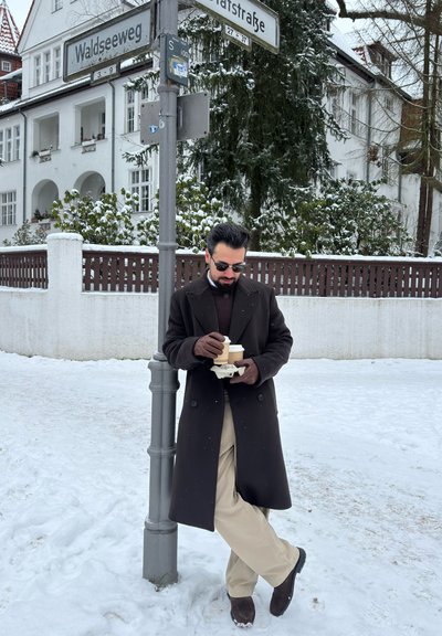 Hombre con abrigo oscuro y guantes sosteniendo dos vasos de café para llevar, apoyado en un poste de señalización en una zona residencial nevada.