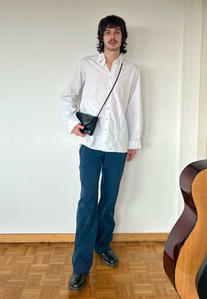 Camisa blanca de botonadura, pantalones azul marino, bolso cruzado negro y zapatos negros. Tejido suave, corte holgado y estilo relajado. Fondo de suelo de madera.